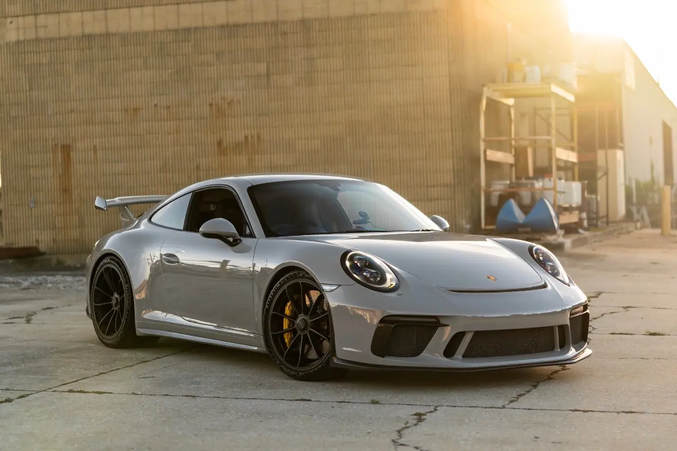 Silver Porsche 911 GT3 sports car with black alloy wheels and rear spoiler, parked on concrete near industrial building in golden hour sunlight.